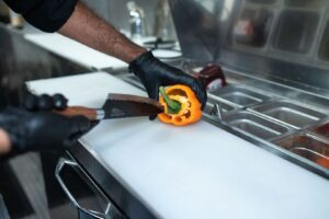 Kock i svarta handskar skär en orange paprika på skärbräda