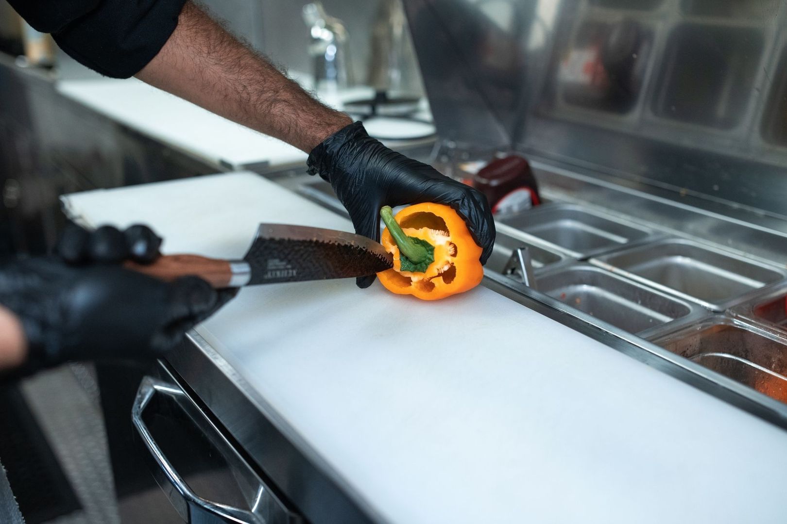 Kock i svarta handskar skär en orange paprika på skärbräda